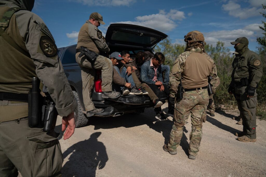 photo of a group of people sitting in the back of an SUV with four border patrol agents around them