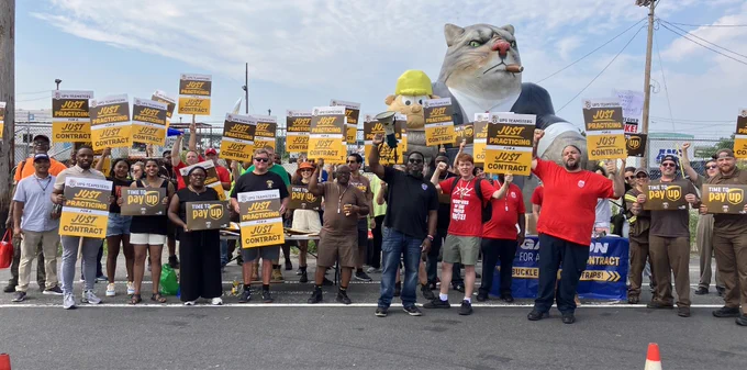 Photo of a large group of people part of a UPS Teamster Practice Picket holding up signs that read "Photo of a group of UPS workers in uniform and holding up signs that say "UPS Teamsters: Just Practicing for a Just Contract" with Scabby in the background