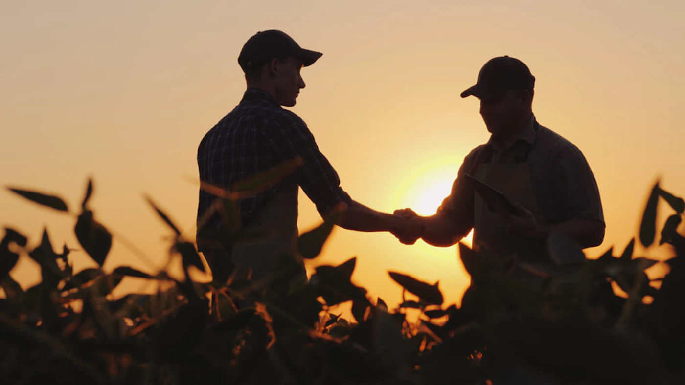 Two people wearing hats who look like farmers shaking hands in a field as the sun sets behind them.
