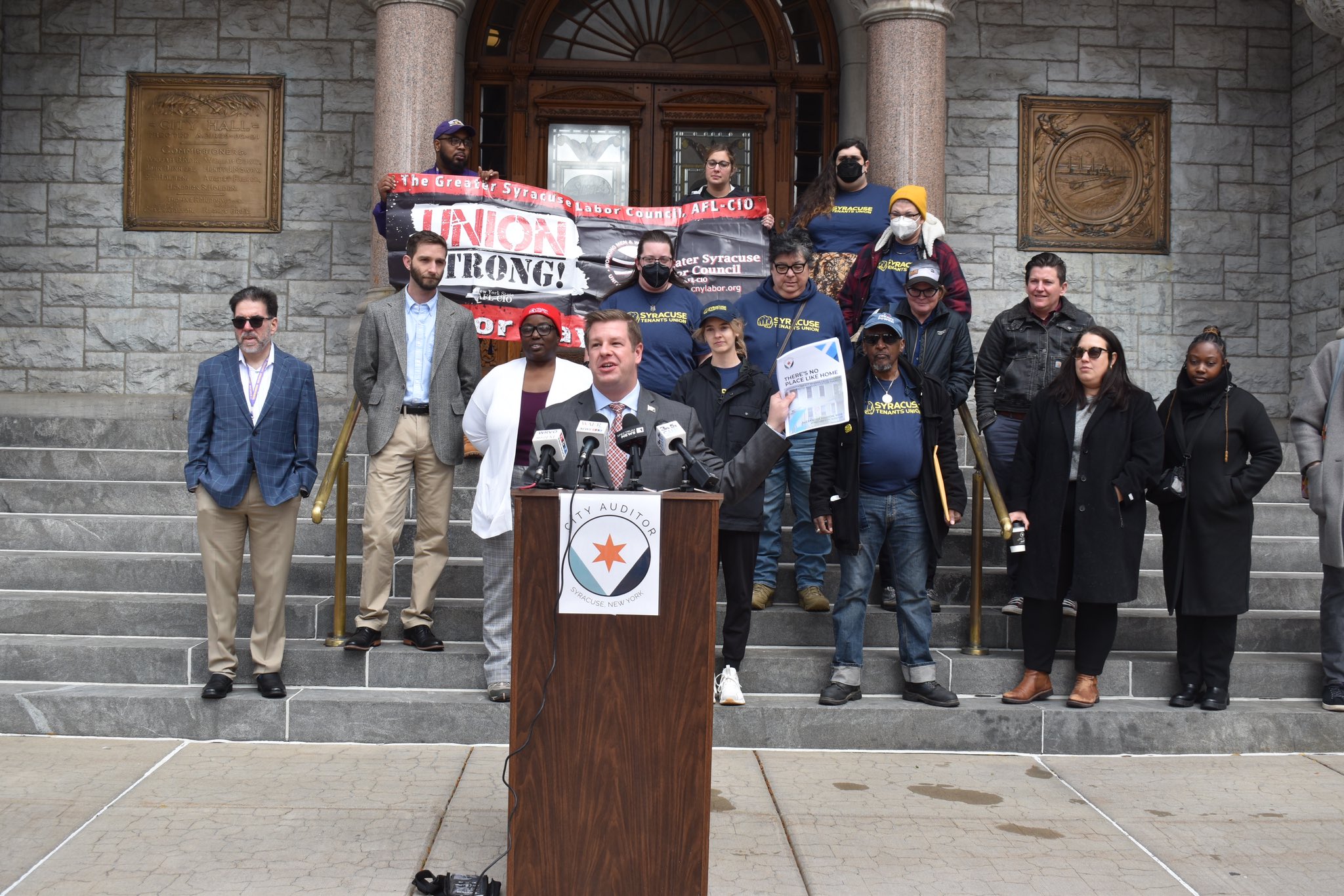 A large group standing around while one person speaks at a podium.