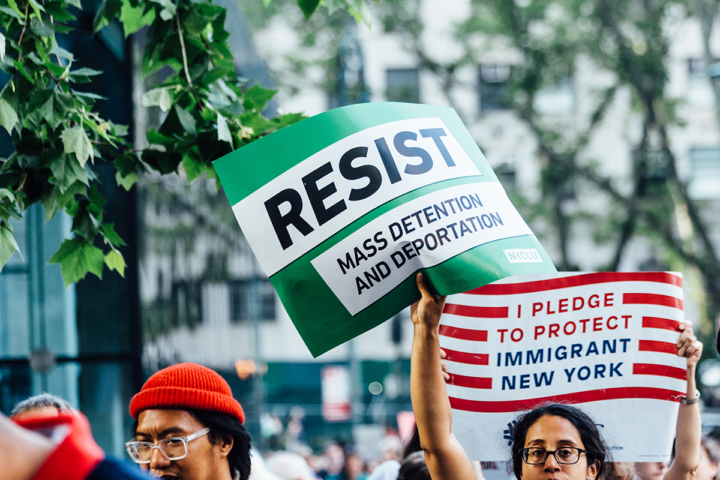 A person holding two signs at a rally. One says "RESIST mass deportation and detention" and the other sign says "I pledge to protect Immigrant New York." Retrieved from the ACLU of New York