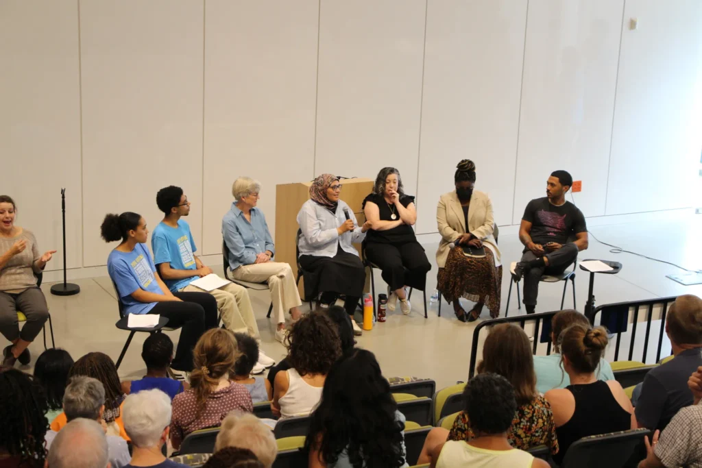 A large panel of speakers speaking to a group of listeners in a large white room.