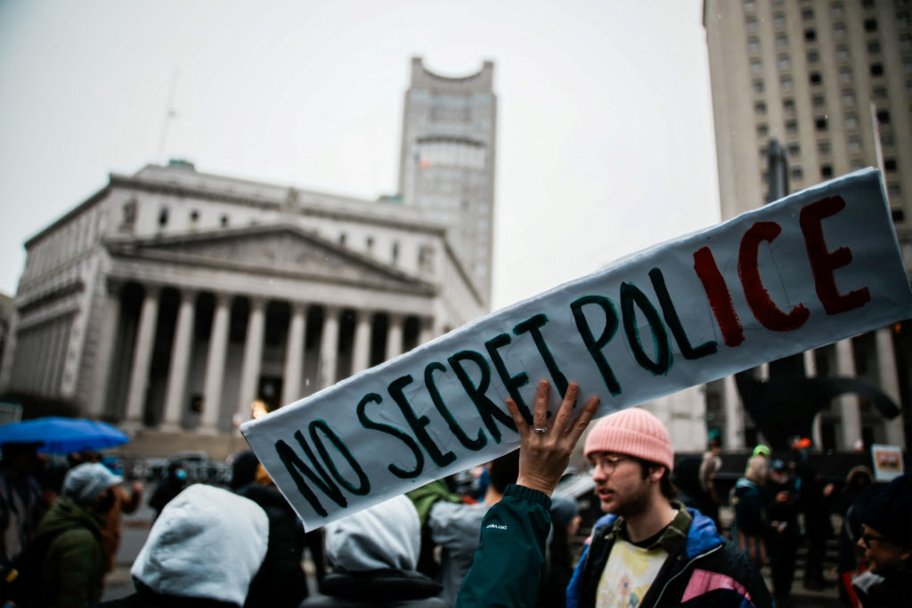 A sign saying "NO SECRET POLICE" held up in front of a government building at a protest. The "ICE" in "POLICE" is in red.